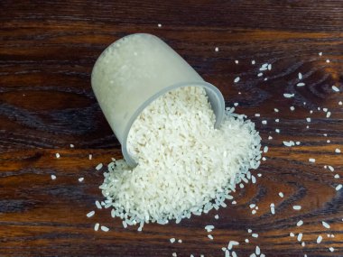 Yakın plan çekimde, temiz bir ölçü kabından koyu, ahşap bir yüzeye dökülen beyaz pirinç sergileniyor. Pirinç taneleri dağılmış, görsel olarak çekici bir düzenleme yapıyorlar.. 