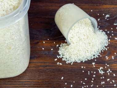 Yakından çekilen beyaz pirinç ölçü kabından koyu kahverengi ahşap bir yüzeye dökülüyor. Solda büyük bir pirinç konteynırı görünür. Kompozisyon iyi ışıklandırılmış yüksek açılı bir çekim..