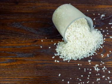 Tepeden çekilen açık bir ölçü kabını yakalar ve zengin desenli, koyu ahşap bir yüzeye beyaz pirinç yığını döker..