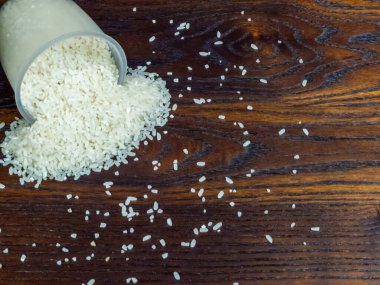Açık plastik bir ölçü kabından koyu ve tahıllı tahta bir yüzeye dökülen pişmemiş beyaz pirincin yakın plan görüntüsü. Pirinç taneleri fincanın etrafına saçılmış. Görsel olarak çekici bir kompozisyon oluşturuyor..