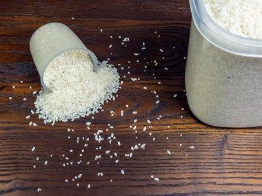 Yakın çekim, beyaz pirincin koyu, ahşap bir yüzeye saçıldığını gösteriyor. Bir ölçü kabı devrildi ve masaya pirinç döküldü. Dökülen taneler için içerik sağlayan pirinç dolu büyük bir konteyner de mevcut..