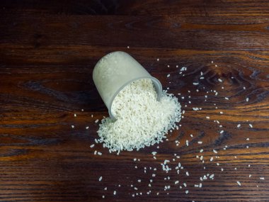 Tepeden aşağı bir atış, devrilmiş temiz bir ölçü kabını yakalar, koyu, ahşap bir yüzeye beyaz pirinç döker. Pirinç, etrafa saçılmış tanelerle küçük bir yığın oluşturur..