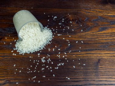 Temiz bir plastik bardağın yakın çekim görüntüsü, pişmemiş beyaz pirinci koyu, parlak ahşap bir yüzeye döküyor. Pirinç fincanın etrafına saçılmış, desenli bir desen oluşturuyor..