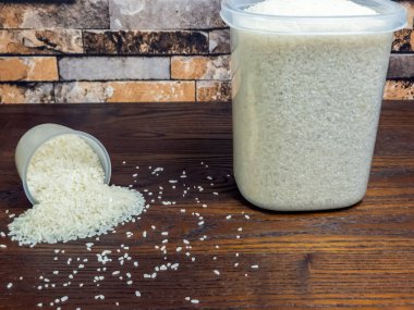 Ahşap bir masanın yakın plan bir resmi, bir ölçü kabı devrilmiş, pirinç taneleri yüzeye dökülüyor. Fincanın yanında içi pirinç dolu büyük bir kap duruyor. Arkaplanda tuğla gibi bir duvar var..