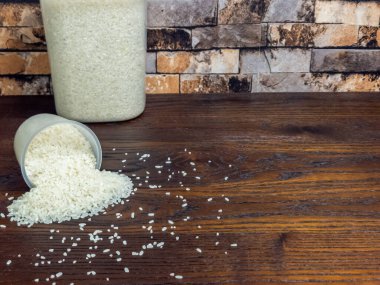 Yakın plan çekimde, koyu ahşap bir yüzeye saçılmış beyaz pirinç yer alır. Bir ölçü kabı yan yatar, pirinci döker, arka plandaki temiz bir kabın içinde daha fazla pirinç görünür. Arka planda desenli tuğla bir duvar var..