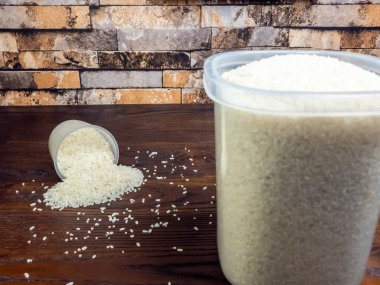 Yakın çekim, devrilmiş bir bardaktan beyaz pirinç döküldüğünü ve koyu kahverengi ahşap bir yüzeyde duran büyük bir kap gösteriyor. Arka planda desenli tuğla bir duvar var. Işıklar parlak ve doğal..