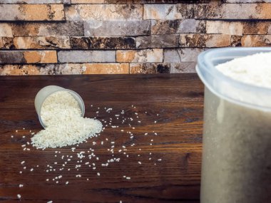 Yakın plan çekimde, ölçüm kabından koyu ahşap bir yüzeye beyaz pirinç dökülüyor. Sağ tarafta pirinçle dolu daha büyük bir kap görülebilir. Arkaplan desenli tuğla bir duvar..