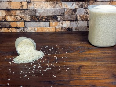 Yakın çekim beyaz pirinç, temiz bir ölçü kabından koyu ahşap bir masaya döküldü. Büyük bir kap pirinç de görülebilir. Arka planda bir tuğla duvar var.. 