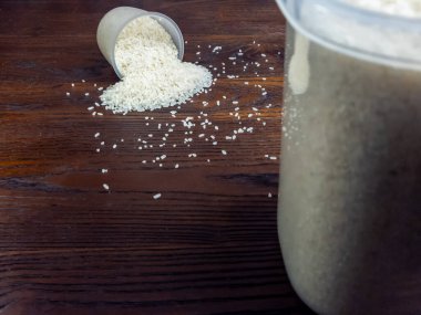 Beyaz pirincin yakın çekim görüntüsü bir ölçü kabından koyu ahşap bir yüzeye dökülüyor. Pirinç veya benzer bir maddeyle dolu büyük bir kap da görülebilir. Görüntü iyi aydınlatılmış, pirinç ve ahşap taneciklerin dokusunu ve detaylarını gösteriyor..