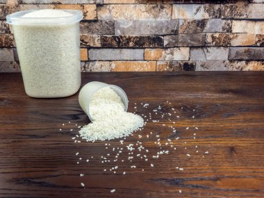 Plastik bardaktan koyu ahşap bir yüzeye dökülen beyaz pirincin yakın plan çekimi. Daha büyük bir kap pirinç fincanın yanında oturur. Arkaplanda tuğla benzeri bir kalıp var..