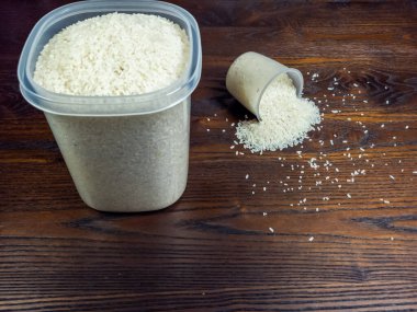 Yakın plan çekimde, eğik bir ölçü kabından dökülen beyaz pirinç ve büyük plastik bir kap, koyu, ahşap bir yüzeye dökülüyor..