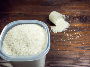 Yakın çekim beyaz pirinç dolu bir konteynırı ve devrilmiş bir ölçü kabını yakalar ve koyu ahşap bir yüzeye pirinç döker. Tahıllar dağılmış, görsel olarak çekici bir bileşim oluşturuyor..