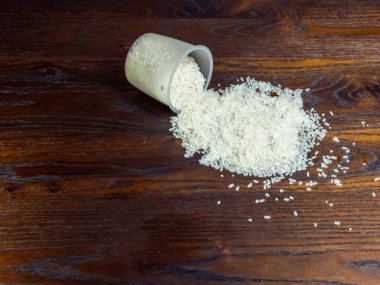 Yakından çekilen bir fotoğraf, zengin, koyu ahşap bir masanın üzerindeki ölçü kabından dökülen pirinci yakalıyor. Kompozisyon, beyaz pirinç ve ahşap dokular arasındaki zıtlığı vurguluyor ve görsel bir ilgi yaratıyor.. 