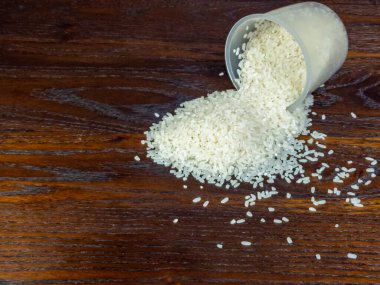 Yakın plan çekimde, temiz bir ölçü kabından çıkan beyaz pirincin koyu ahşap bir yüzeye döküldüğünü görüyoruz..