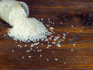 Yakından çekilmiş, pişmemiş beyaz pirinçler temiz bir plastik ölçü kabından koyu renkli bir tahta yüzeye dökülüyor. Bileşim, pirinç ve ahşap taneciklerin dokusunu vurguluyor ve görsel olarak çekici bir kontrast oluşturuyor..
