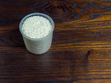 Yakın plan çekimde, koyu ahşap bir yüzeye yerleştirilmiş pişmemiş beyaz pirinçle dolu plastik bir ölçü kabı yer alıyor. Görüntü yüksek açılı bir perspektiften çekiliyor, pirinç ve ahşap taneciklerin dokusunu vurguluyor..