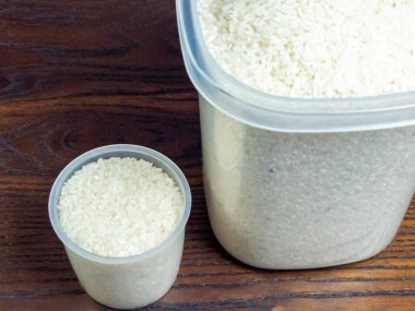 Plastik bir kabın içinde yakın plan pirinç taneleri ve ölçü kabı. Konteynır kısmen pirinçle dolu ve kap ağzına kadar dolu. Arkaplan, doğal ve sıcak bir estetik sunan ahşap bir yüzey..