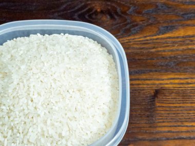 Yakın çekim bir vitrin içinde pişmemiş beyaz pirinç bulunan plastik bir kap. Konteynır koyu ahşap bir yüzeyde duruyor ve zıt bir zıt zemin oluşturuyor. Görüntü pirinç tanelerinin dokusunu ve detaylarını vurgular..