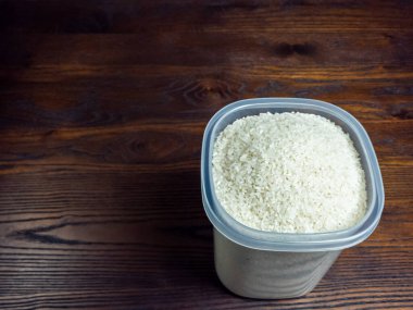 Beyaz pirinçle dolu plastik bir konteynırın yakın plan çekimi. Koyu, ahşap bir arka plan üzerine kurulmuş..