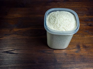 Yakından çekilen bir fotoğrafta, pişmemiş beyaz pirinçle dolu temiz bir plastik kap sergileniyor. Konteynır koyu, ahşap bir yüzeye yerleştiriliyor..