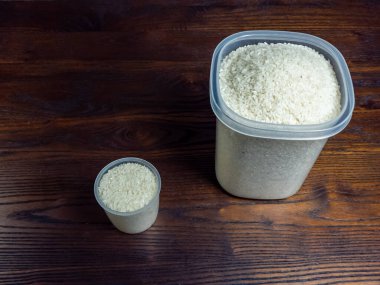 Yüksek açılı, yakın planda bir fotoğraf büyük bir plastik kap ve daha küçük bir kap, her ikisi de pişmemiş beyaz pirinçle dolu, koyu ahşap bir yüzeye yerleştirilmiş. Işıklandırma doğal, pirinç ve ahşap taneciklerin dokusunu vurguluyor..