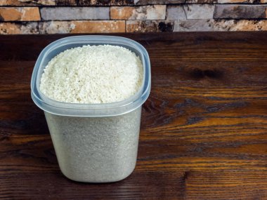 Pişmemiş beyaz pirinçle dolu şeffaf bir plastik kap, siyah bir ahşap masada oturur. Arka planda tuğla bir duvar var. Görüntü iyi aydınlatılmış, pirincin dokusuna ve pirinç ile masa arasındaki kontrast üzerine odaklanmış..