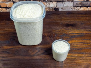 Bir fotoğraf, biri büyük diğeri küçük olmak üzere siyah tahta bir yüzeyde beyaz pirinçle dolu iki plastik kap gösteriyor. Pirinç her iki konteynırda da görülebilir ve bileşimi boyut olarak kontrastı vurguluyor. 