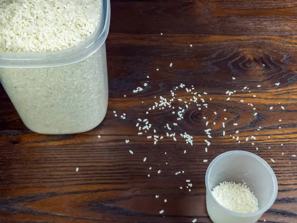 Pirinç dolu temiz bir kap ile ahşap bir masa yakın çekim, kısmen pirinç dolu bir ölçü kabı, ve yüzey boyunca yayılmış pirinç.