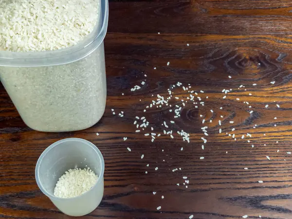 Yüksek açılı çekim vitrinleri zengin, koyu ahşap bir yüzeye pirinç taneleri saçar. Pirinçle dolu büyük bir kap çerçevede kısmen yer alır, küçük bir ölçü kabıyla birlikte pirinç de içerir..