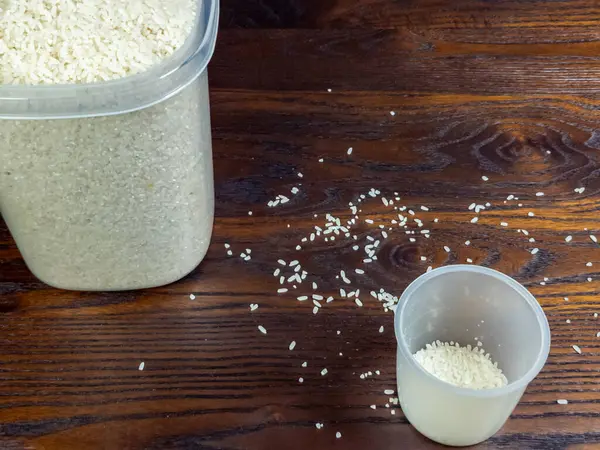 Yakın plan çekimde, koyu ahşap bir yüzeyde dağınık tanecikli, pişmemiş beyaz pirinç konteynırı yer alıyor. Bir ölçü kabında pirincin bir kısmı bulunur. Aydınlatma ve kompozisyon tanelerin dokusunu ve detaylarını vurgular.