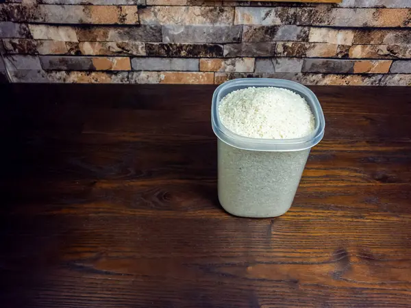 Yakın plan çekimde, koyu ahşap bir yüzeye yerleştirilmiş beyaz pirinçle dolup taşan plastik bir kap görülüyor. Arka planda bir tuğla duvar, doku ve derinlik ekleniyor. Işık, pirincin dokusunu ve renk kontrastını vurguluyor..