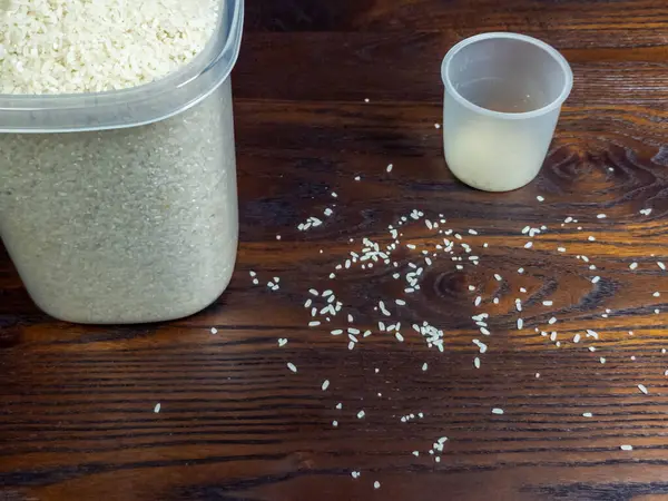 Tepeden tırnağa bir çekimde ölçü kabının yanında beyaz pirinç konteynırı sergilenir. Dağıtılmış pirinç taneleri zengin kahverengi ahşap yüzey.