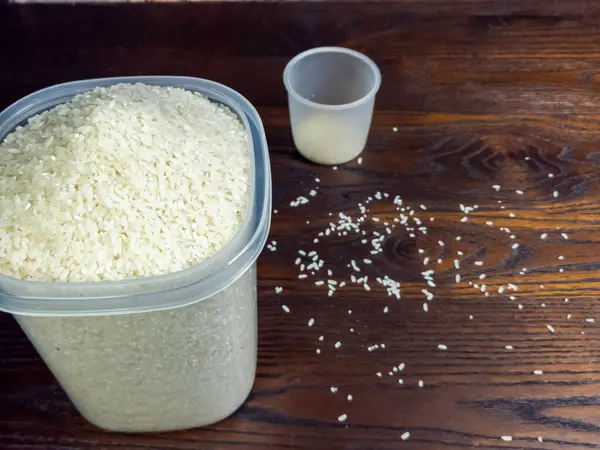 Pişmemiş beyaz pirinçle dolu bir konteynırın yakın çekim görüntüsü, yanında bir ölçü kabı ve ahşap bir yüzeye saçılmış taneler. Görüntü pilavın dokusunu ve rengini vurguluyor, özellikle de yemek ve yemek hazırlama konularında.. 