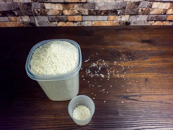 Yakın plan çekimde beyaz pirinçle dolup taşan büyük bir konteynır görülüyor. Yanında aynı pirinçle dolu küçük bir ölçü kabı var. Sahne koyu, ahşap bir yüzeye kurulmuş, dağınık taneciklerle, tuğladan yapılmış bir arka plana karşı..