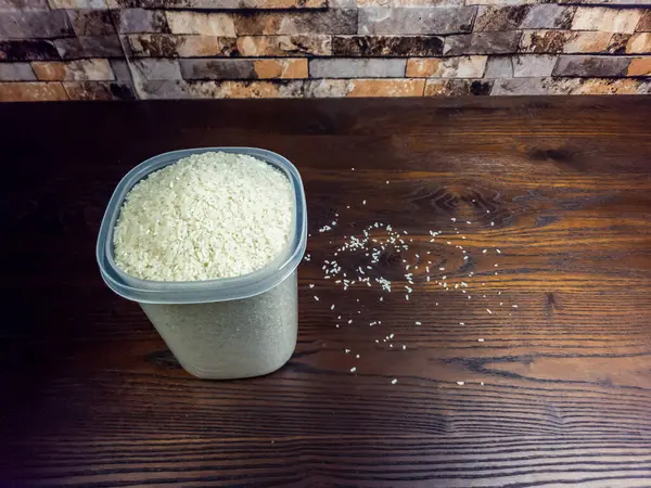 Yakın plan çekimde, beyaz pirinçle dolu, siyah ahşap bir yüzey üzerinde duran şeffaf plastik bir kap sergileniyor. Masada dağınık pirinç taneleri de mevcuttur. Arka planda desenli tuğla bir duvar var..