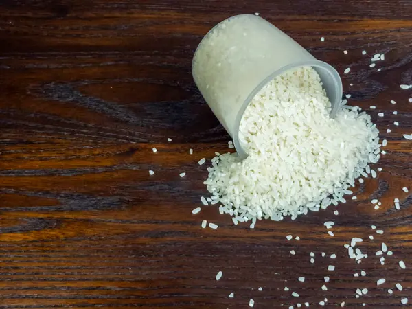 Tepeden çekilen açık bir ölçü kabını yakalar ve zengin desenli, koyu ahşap bir yüzeye beyaz pirinç yığını döker..