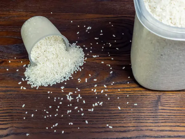 Yakın çekim, beyaz pirincin koyu, ahşap bir yüzeye saçıldığını gösteriyor. Bir ölçü kabı devrildi ve masaya pirinç döküldü. Dökülen taneler için içerik sağlayan pirinç dolu büyük bir konteyner de mevcut..