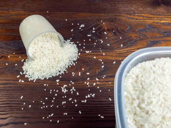 Plastik bir bardaktan siyah bir tahta yüzeye dökülen beyaz pirincin yakın plan çekimi. Bazı taneler fincanın etrafına saçılmış ve sağ alt köşede bir kap pirinç görünüyor.. 