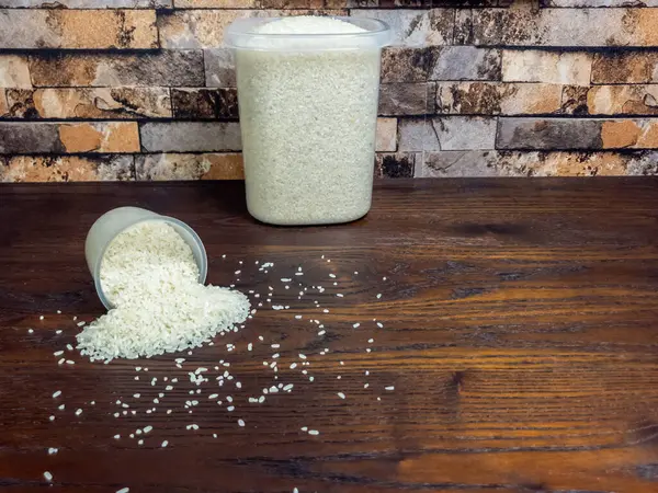 Ahşap bir masada pirinç ve konteynırların yer aldığı durgun bir yaşam görüntüsü. Temiz bir plastik bardak devrildi ve tahta yüzeye beyaz pirinç döküldü. Arkasında pirinç dolu daha büyük bir konteynır duruyor..