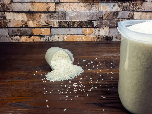 Yakından çekilen beyaz pirinç ölçü kabından koyu ahşap bir yüzeye dökülüyor. Yakınlarda büyük bir pirinç konteynırı var. Arka planda bir tuğla duvar ve doku var. Görüntü bir bolluk ve hazırlık hissi taşır..