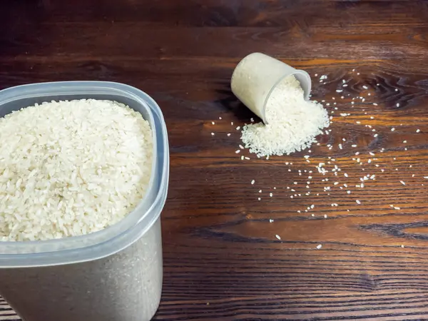 Yakın çekimde beyaz pirinç, büyük bir kap pirinç ve pirinç dökülmüş bir ölçü kabı sergilenir. Pirinç taneleri ahşap yüzeye saçıldı. Görüntü gıda hazırlığını vurguluyor.