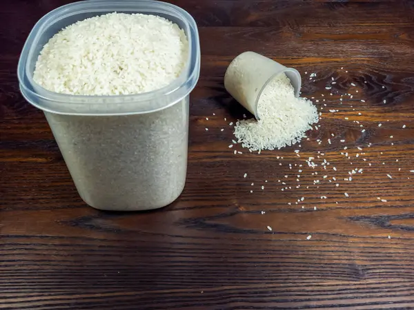 Yakın plan çekimde, eğik bir ölçü kabından dökülen beyaz pirinç ve büyük plastik bir kap, koyu, ahşap bir yüzeye dökülüyor..