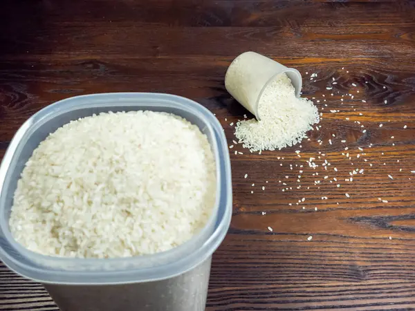 Yakın çekim beyaz pirinç dolu bir konteynırı ve devrilmiş bir ölçü kabını yakalar ve koyu ahşap bir yüzeye pirinç döker. Tahıllar dağılmış, görsel olarak çekici bir bileşim oluşturuyor..