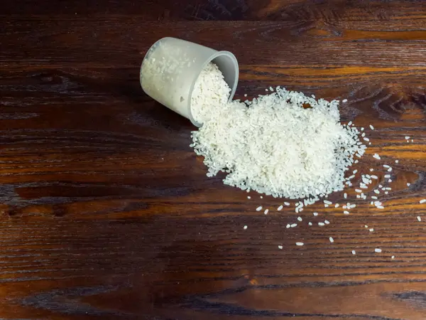 Yakından çekilen bir fotoğraf, zengin, koyu ahşap bir masanın üzerindeki ölçü kabından dökülen pirinci yakalıyor. Kompozisyon, beyaz pirinç ve ahşap dokular arasındaki zıtlığı vurguluyor ve görsel bir ilgi yaratıyor.. 