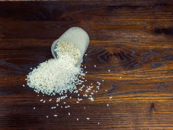 Yakın çekim, üzerinde beyaz pirinci koyu kahverengi bir ahşap yüzeye döken açık bir ölçüm kabını içerir. Tahta tohumu görünür, doku ve görsel ilgi ekliyor..