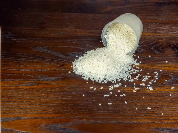 Yakın plan çekimde, saydam bir ölçüm kabından dökülen beyaz pirincin zengin, koyu bir tahta yüzeye döküldüğü görülüyor. Bileşim, pirinç tanelerinin ahşap taneciklerine karşı dokusunu ve kontrastını vurguluyor..