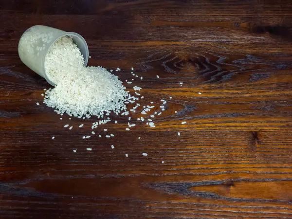 Bir stüdyo, şeffaf bir konteynırdan koyu kahverengi bir ahşap yüzeye dökülen beyaz pirinci sergiliyor. Görüntü hem pirincin hem de tahtanın dokusunu vurgular, görsel olarak çekici bir bileşim yaratır..