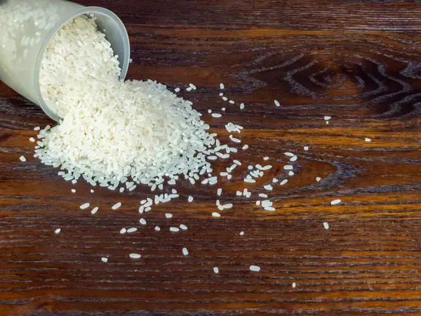 Yakından çekilmiş, pişmemiş beyaz pirinçler temiz bir plastik ölçü kabından koyu renkli bir tahta yüzeye dökülüyor. Bileşim, pirinç ve ahşap taneciklerin dokusunu vurguluyor ve görsel olarak çekici bir kontrast oluşturuyor..