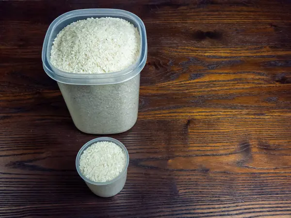 Üstünde pişmemiş beyaz pirinçle dolu iki şeffaf plastik kap olan tepeden tırnağa bir çekim. Konteynırlar koyu, bol tahıllı ahşap bir yüzeye yerleştirilmiş ve sağ tarafında geniş bir kopya alanı var..