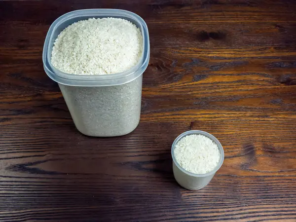 Biri büyük diğeri küçük olmak üzere beyaz pirinçle dolu iki plastik kap, koyu ahşap bir masanın üzerinde duruyor. Görüntü pirincin dokusunu ve rengini ve konteynırlarla ahşap arasındaki kontrastı vurguluyor..